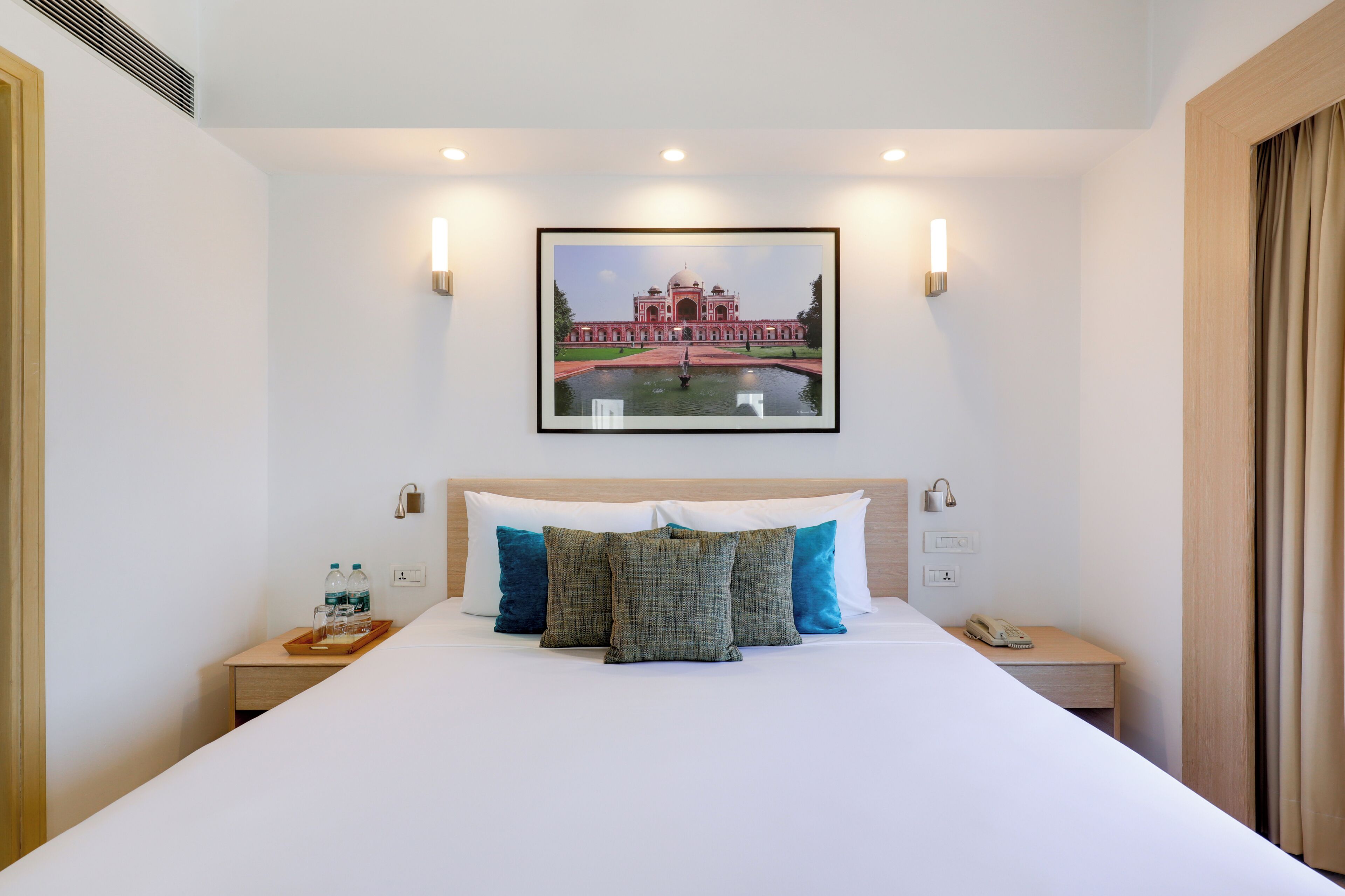 Habitación de hotel con una cama grande, un cuadro enmarco de un edificio, lámparas de noche, una mesita de noche con un teléfono y una bandeja con botellas.