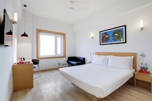 Habitación de hotel con cama, escritorio, silla, mesita, ventilador de techo, ventana y un cuadro en la pared.