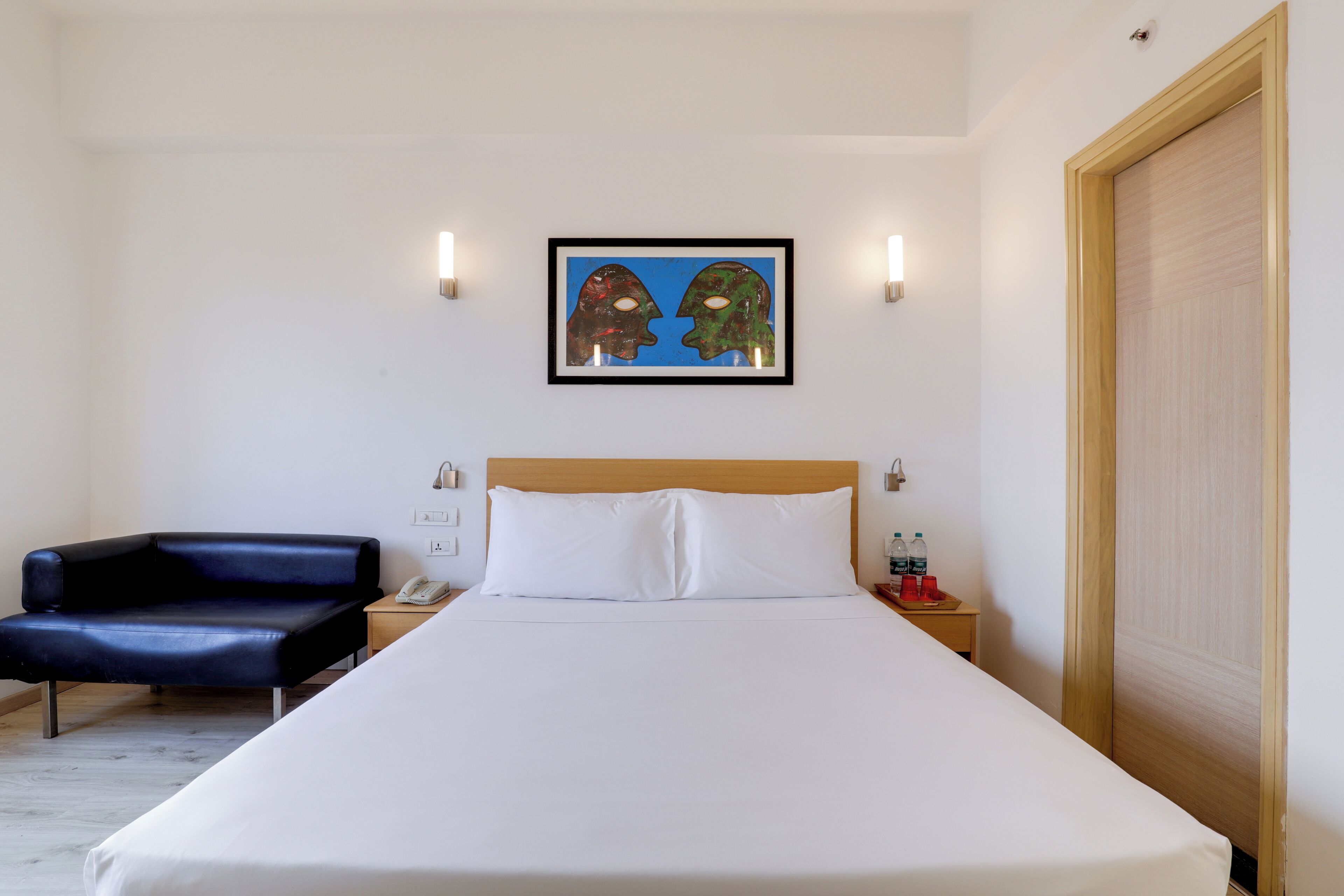 A hotel room with a bed, a black leather chair, a bedside table with a phone and a bottle, and a painting on the wall.