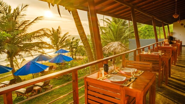 A restaurant with wooden tables and chairs, blue umbrellas, and a view of the ocean and palm trees. - The Seascape (Matara)