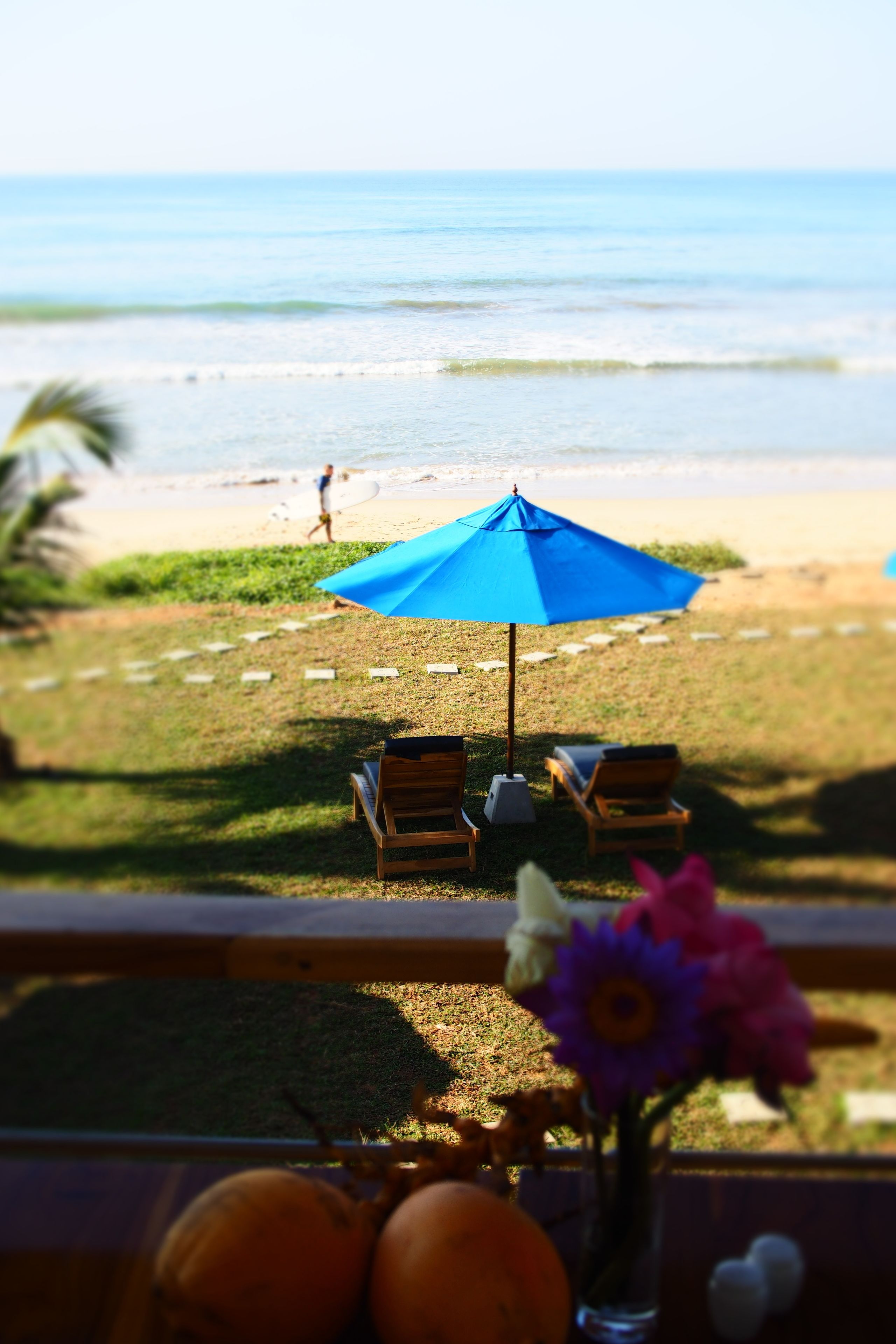 A beach view with a blue umbrella, lounge chairs, and a vase with flowers.