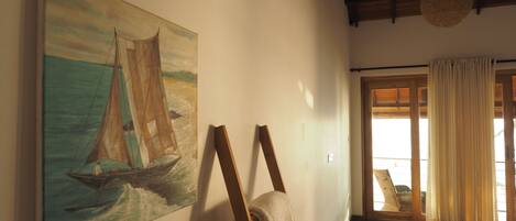 Une pièce avec une échelle en bois appuyée contre un mur, un tableau représentant un voilier et, vue à travers une porte vitrée, un espace extérieur avec une chaise.