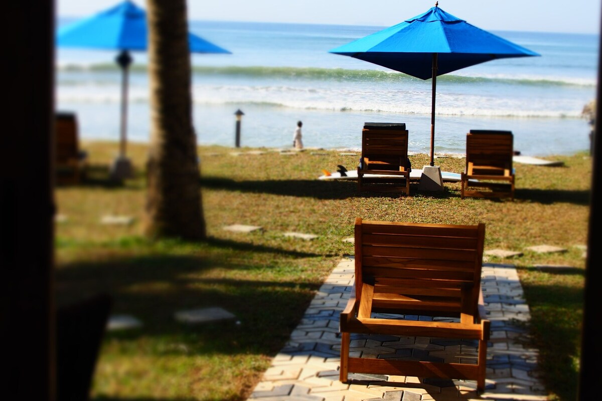Un patio frente a la playa con sillas de madera y una sombrilla azul, con vistas al océano.