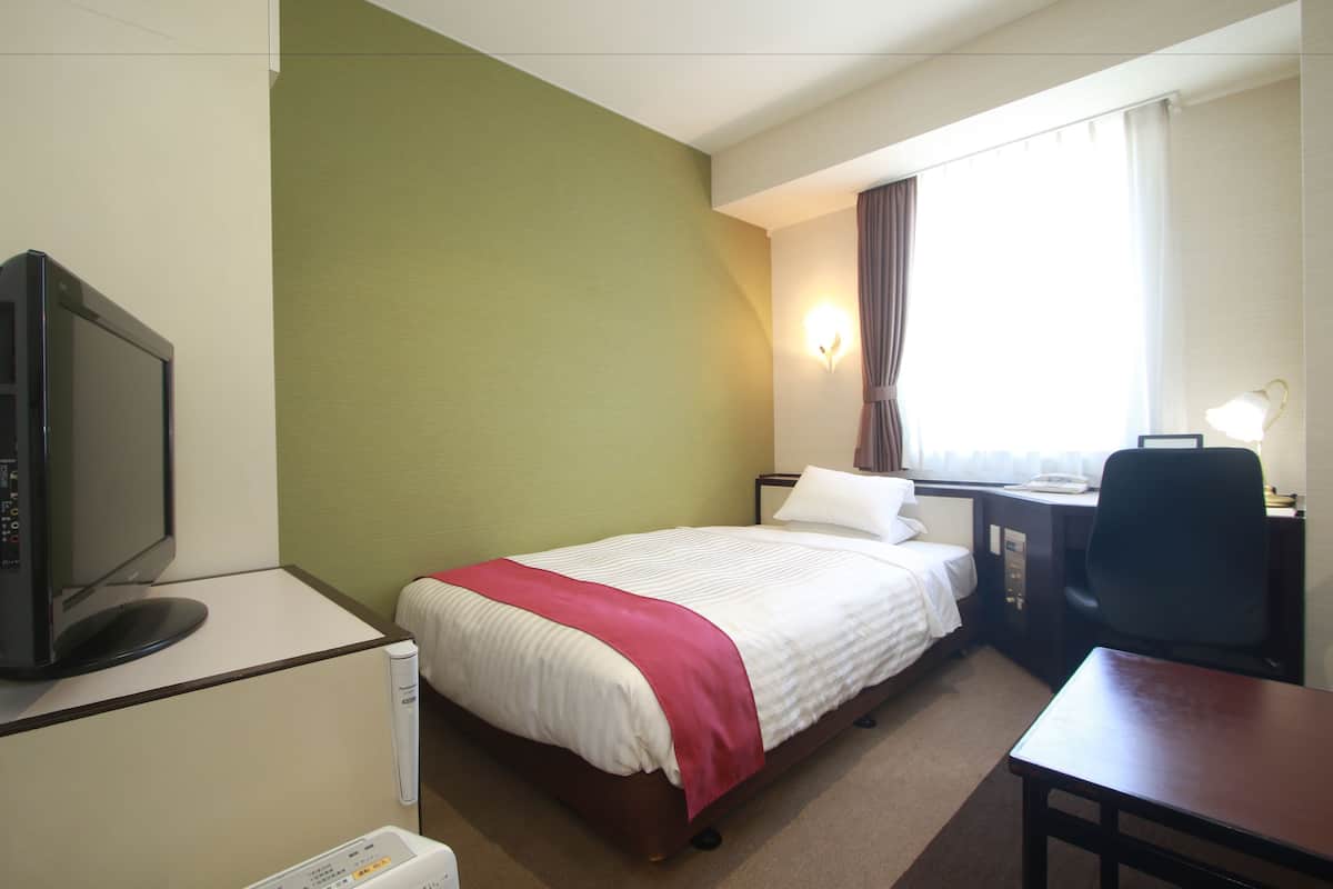 Una habitación de hotel con cama, escritorio, silla, televisor y ventana con cortinas.