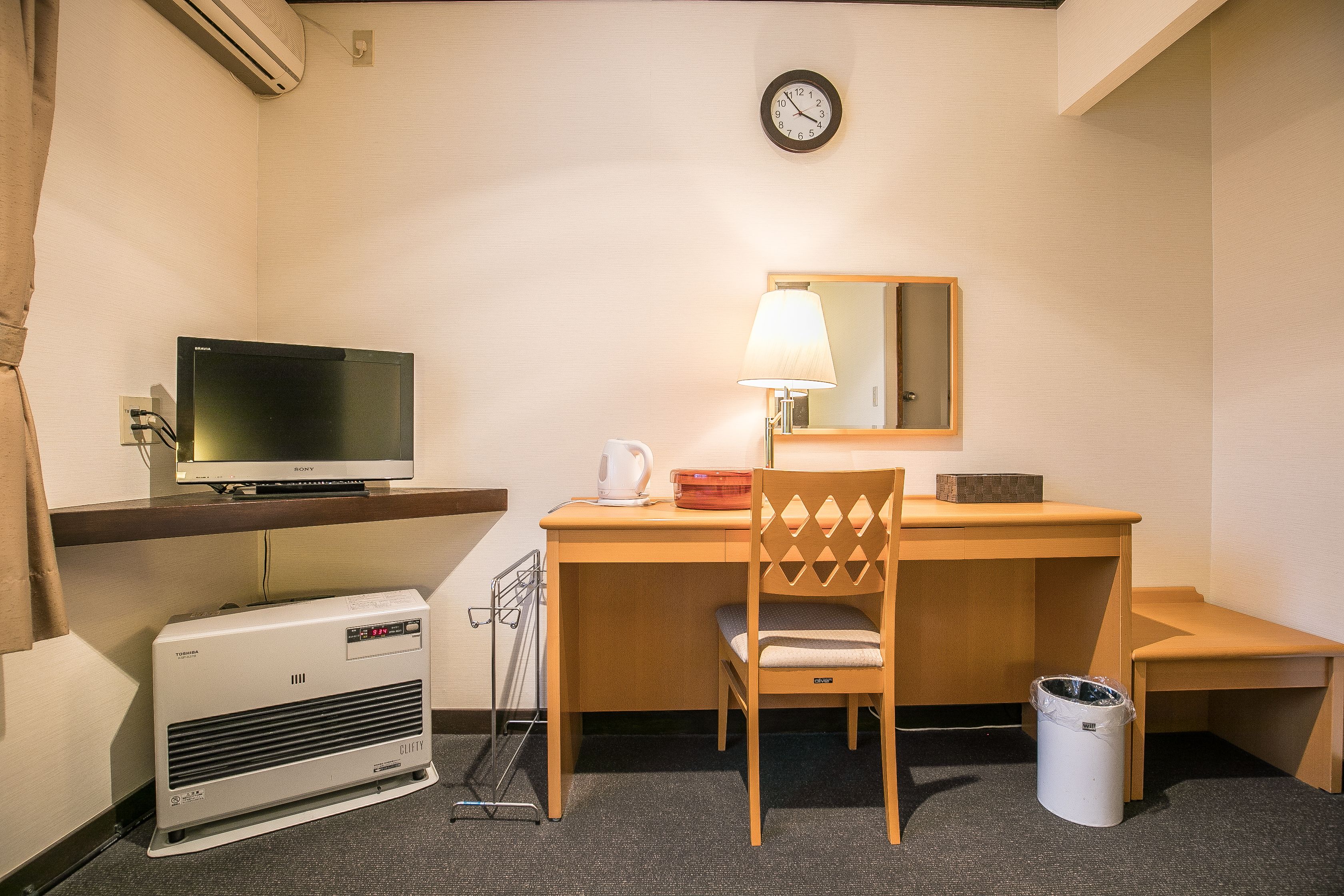 A hotel room with a desk, chair, TV, lamp, clock, and air conditioning unit.