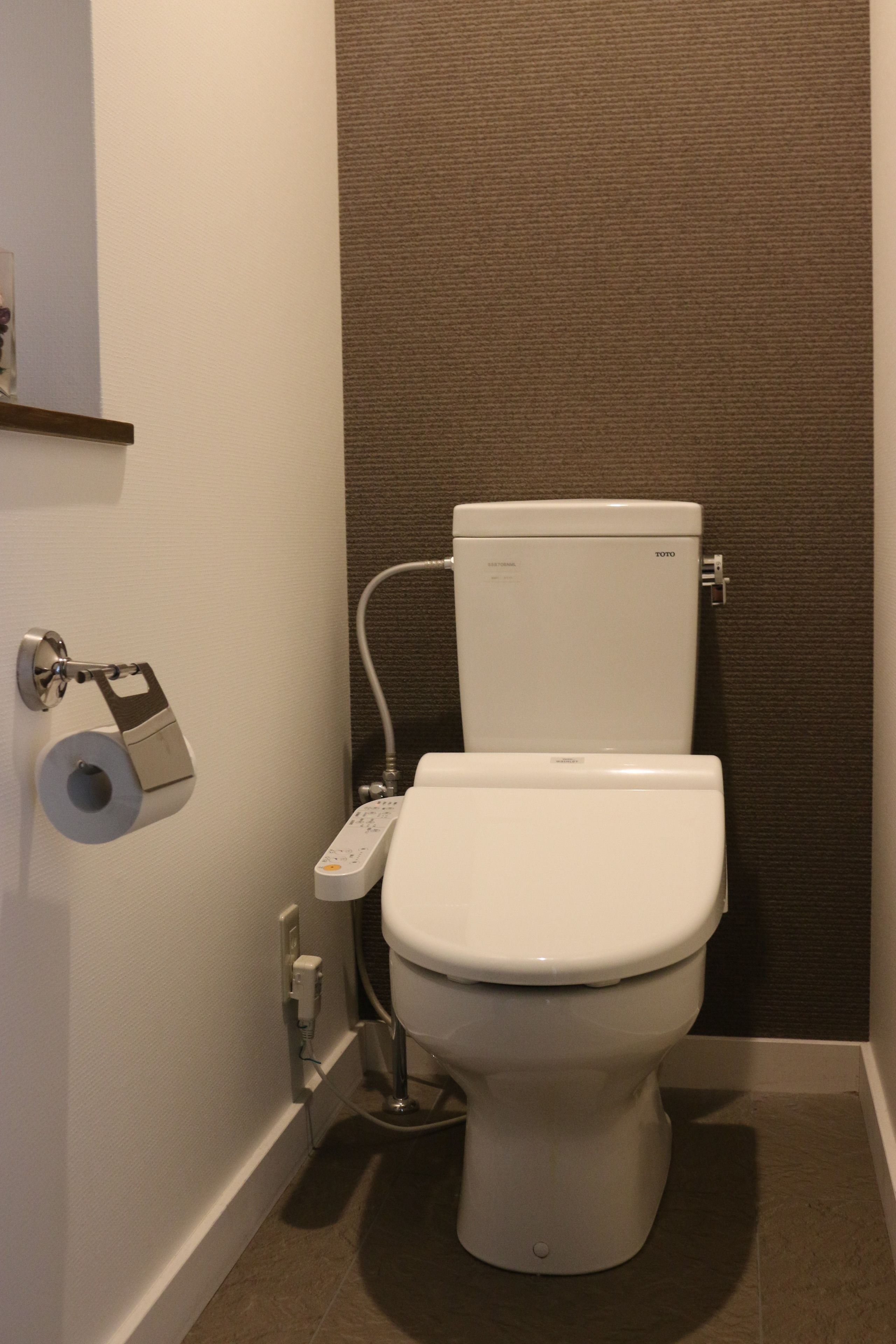 Inodoro blanco estándar con panel de control, dispensador de papel higiénico y pulsador de descarga en la pared.