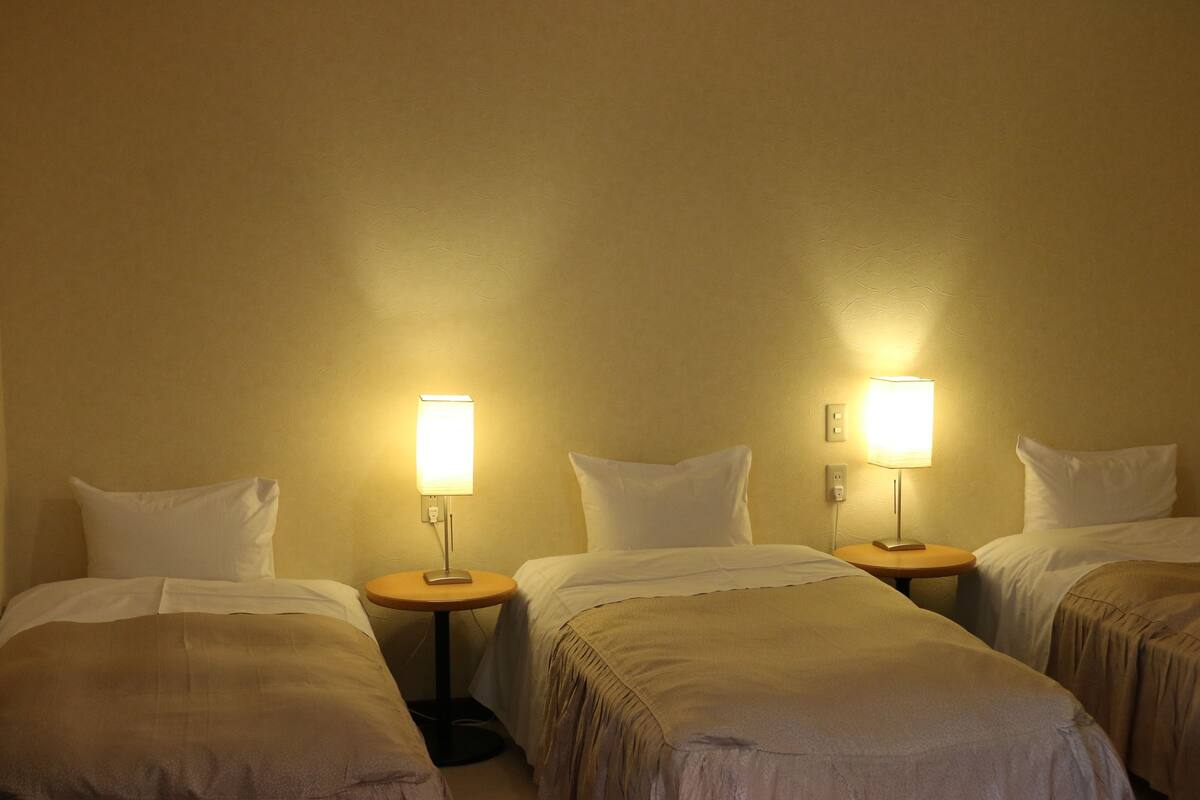 Ein Hotelzimmer mit drei Einzelbetten, jeweils mit Nachttisch und Lampe.