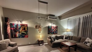 Een woonkamer met een bank, salontafel en vloerkleed. Er hangen twee schilderijen aan de muur, een lamp en een airconditioner.