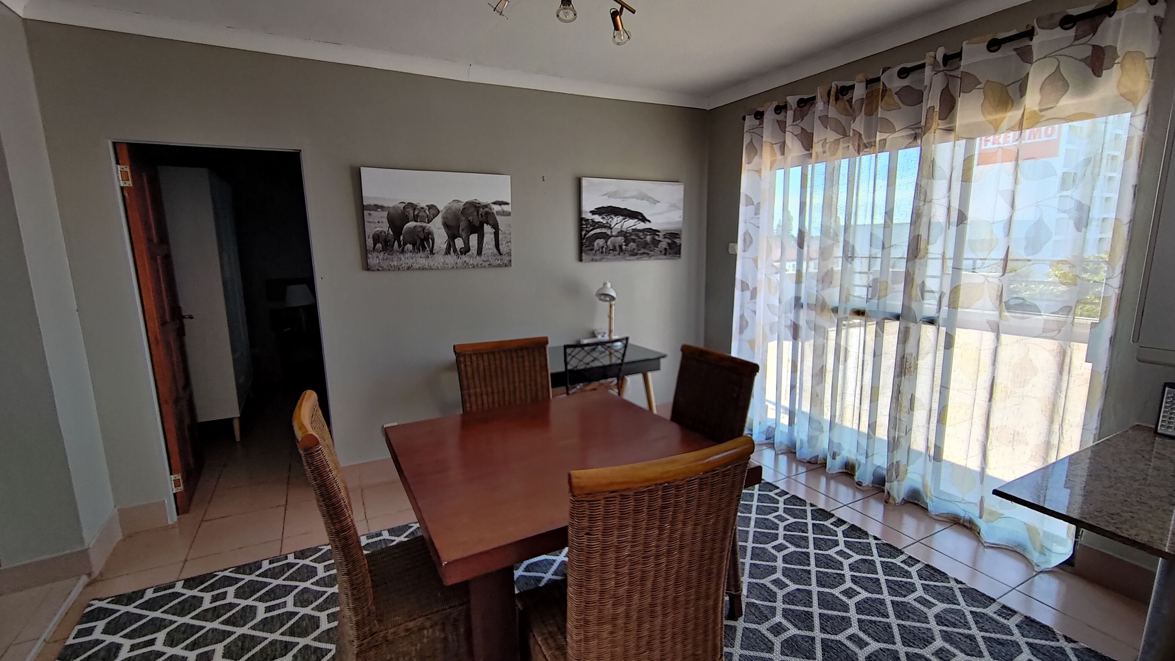 Een eetgedeelte met een houten tafel, vier rieten stoelen, twee aan de muur hangende afbeeldingen en een kamerbreed tapijt met een patroon.