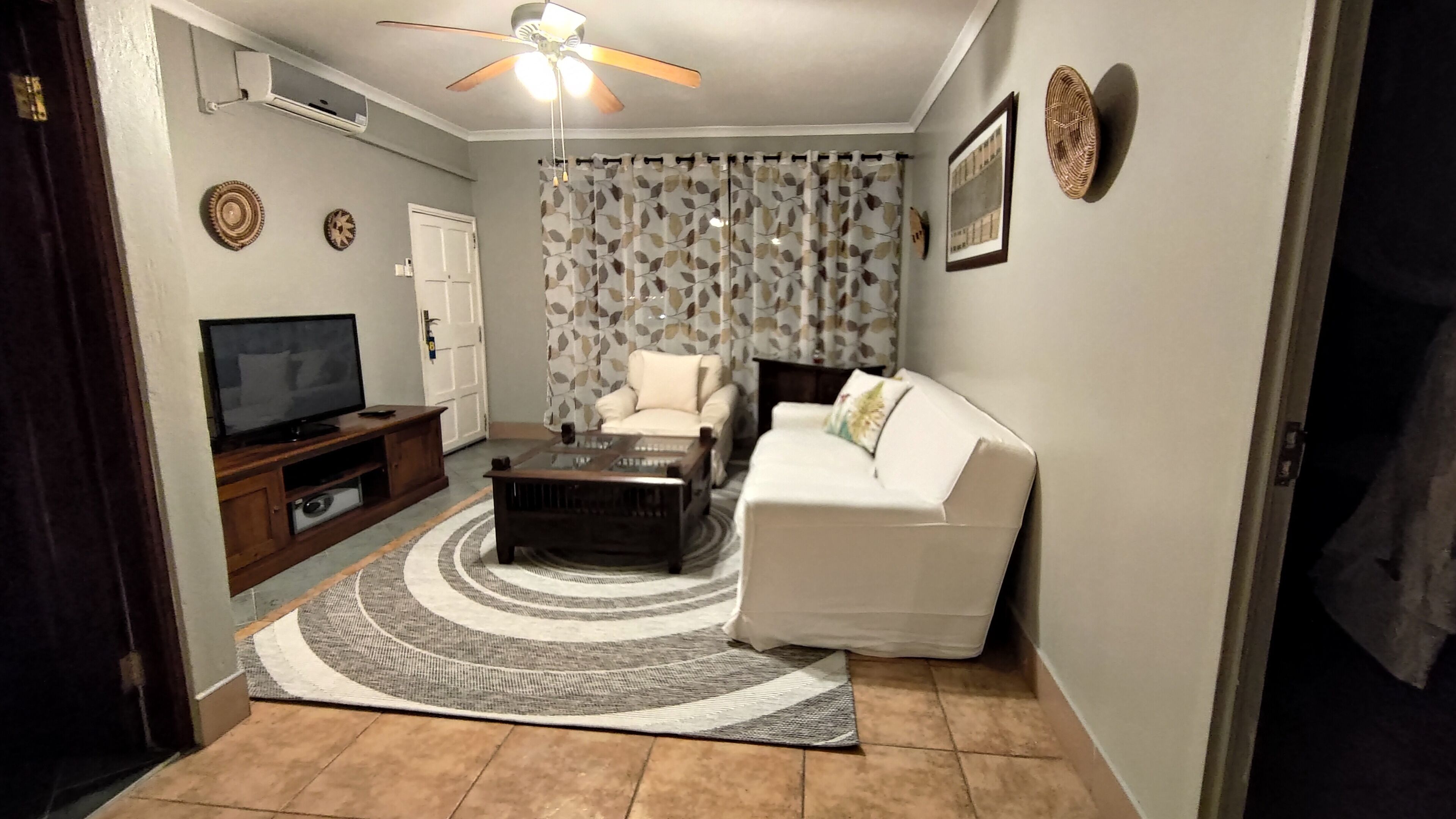 A living room with a TV, sofa, armchair, coffee table, patterned curtains, and a ceiling fan.