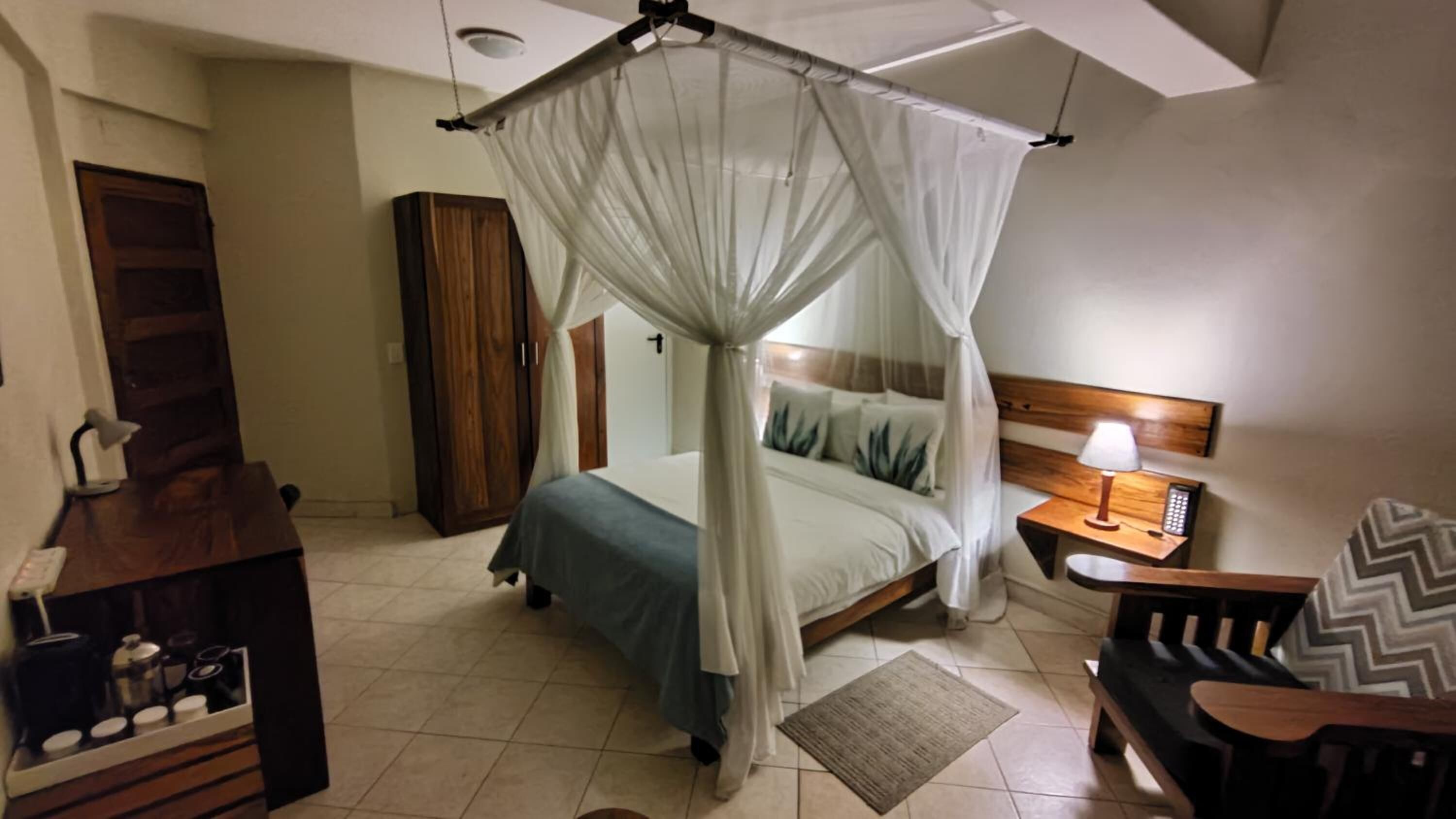 Un dormitorio con cama a dosel, una mesita de noche con lámpara, un armario de madera y una pequeña mesa con cafetera.
