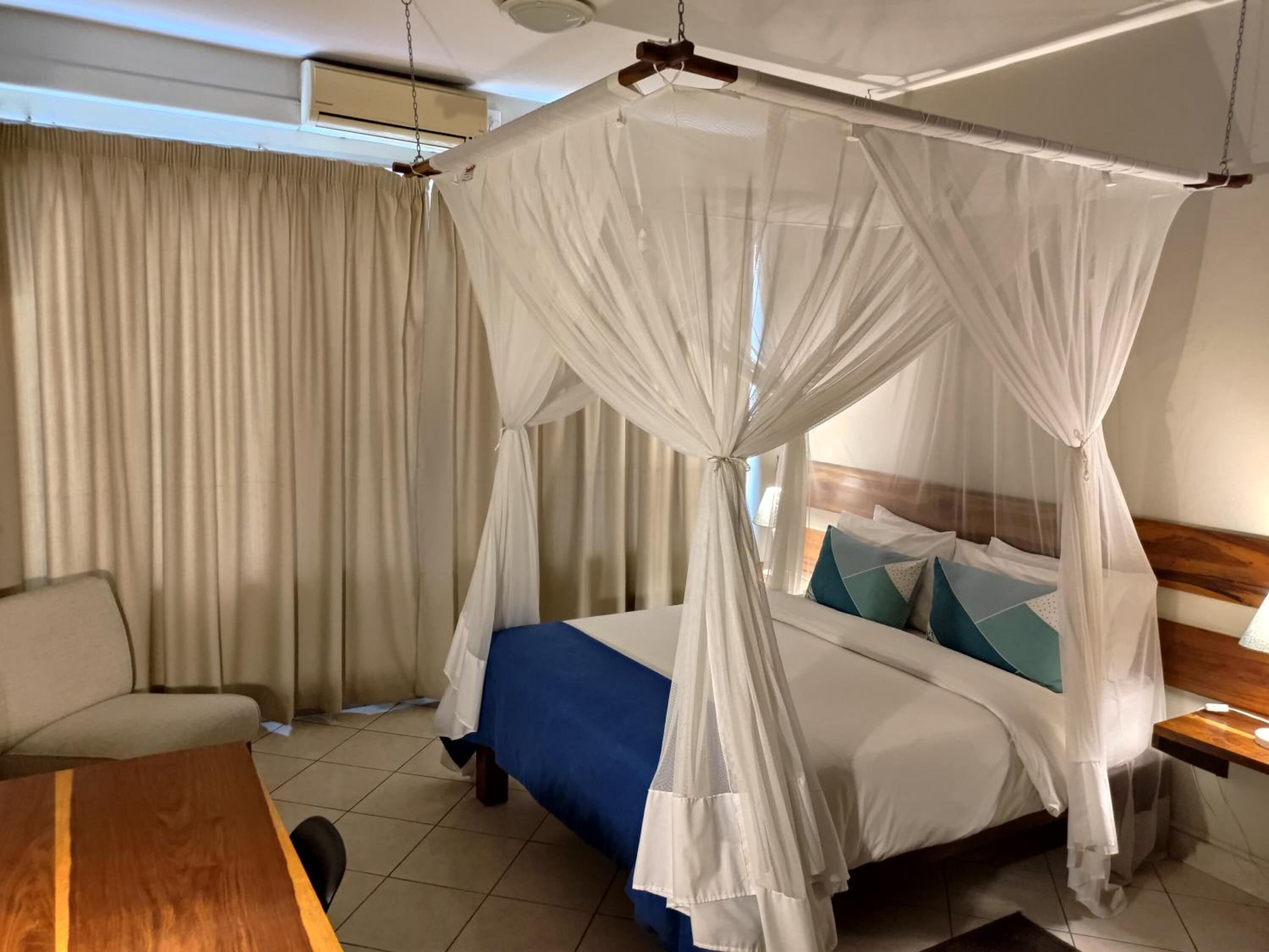 A hotel room with a bed under a canopy, a sofa, a wooden table, and a window with curtains.