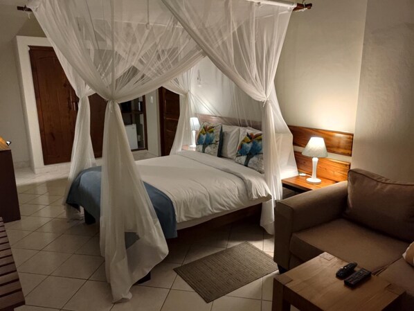 A bedroom with a four-poster canopy bed, a bedside table with a lamp, a sofa, and a small table with two remote controls.