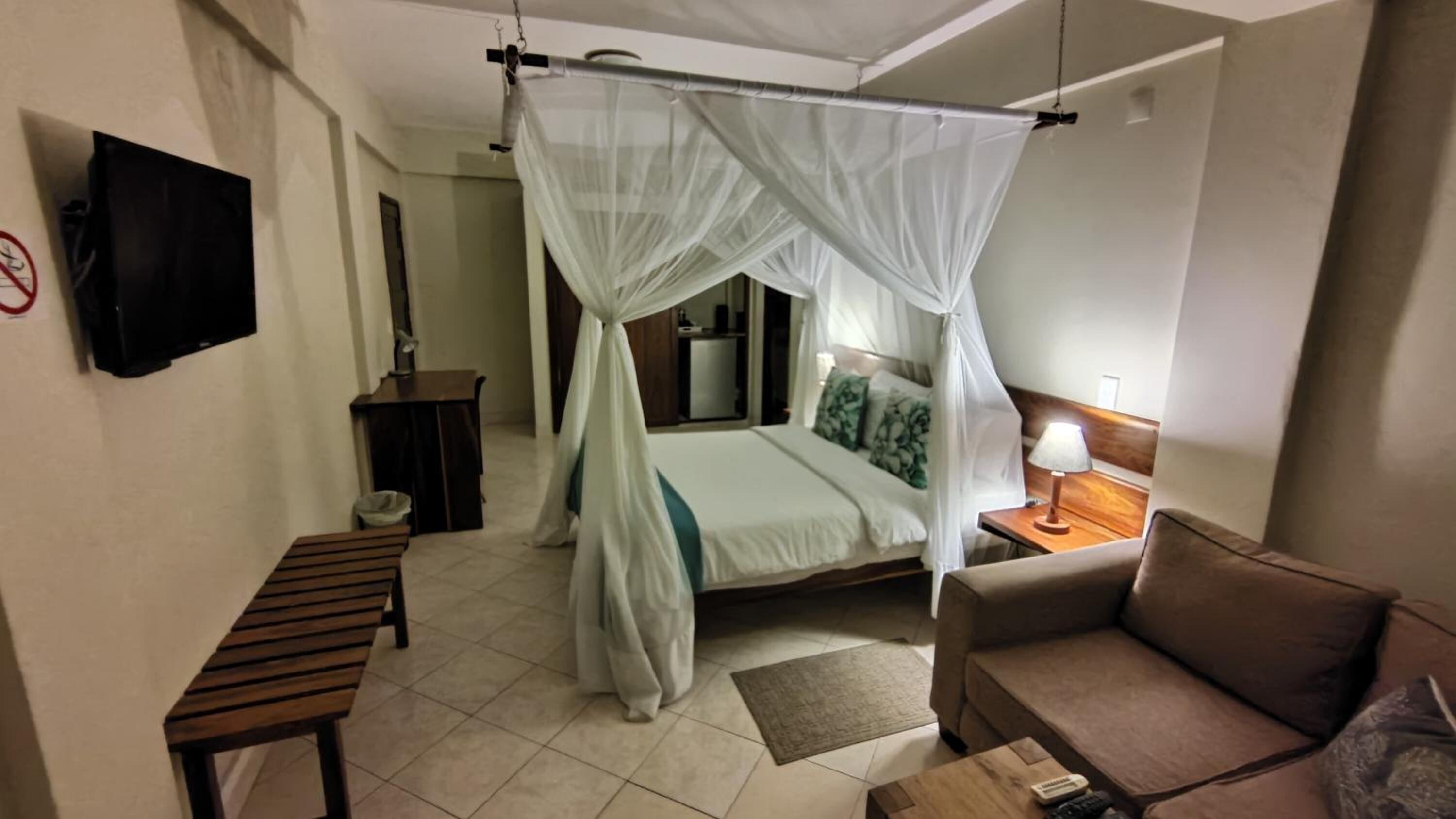 Habitación de hotel con una cama bajo dosel, un sofá, una mesita, un televisor montado en la pared y un escritorio con una silla.