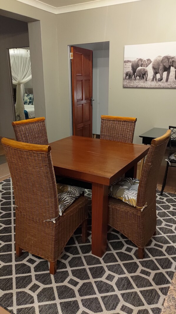 Un comedor con una mesa de madera y sillas de mimbre, un tapete estampado y una puerta que da a otra habitación.