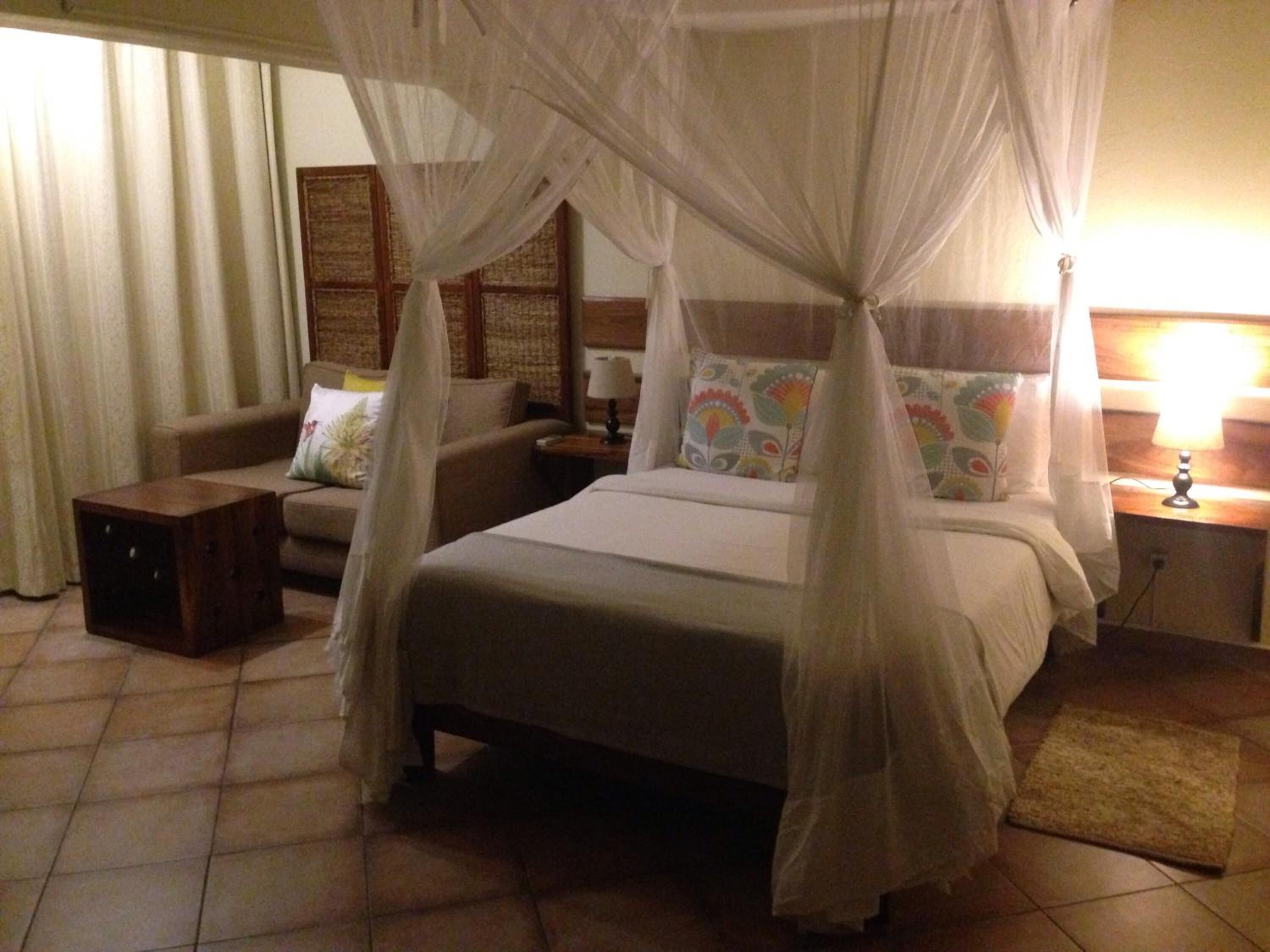 A bedroom with a four-poster bed, a nightstand with a lamp, a small wooden side table, and a sofa with patterned cushions.