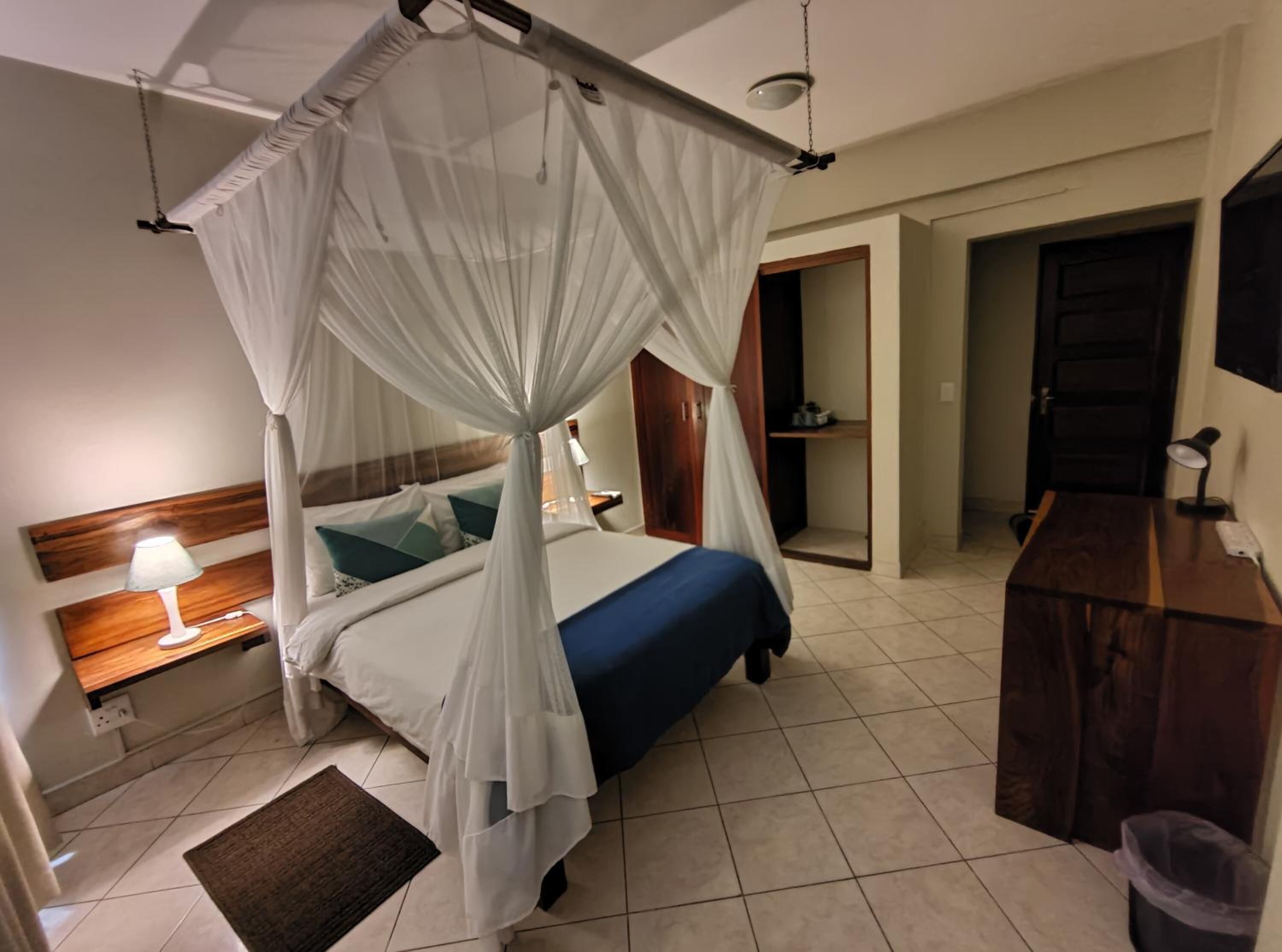 A bedroom with a four-poster bed, a bedside table with a lamp, a wooden dresser, and a door leading to another room.