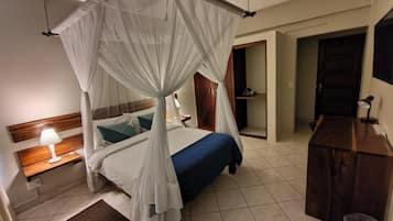 A bedroom with a four-poster bed, a bedside table with a lamp, a wooden dresser, and a door leading to another room.