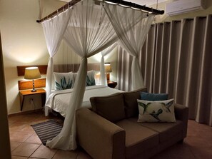 A hotel room with a four-poster bed, a sofa, two bedside tables with lamps, and a large window with curtains.