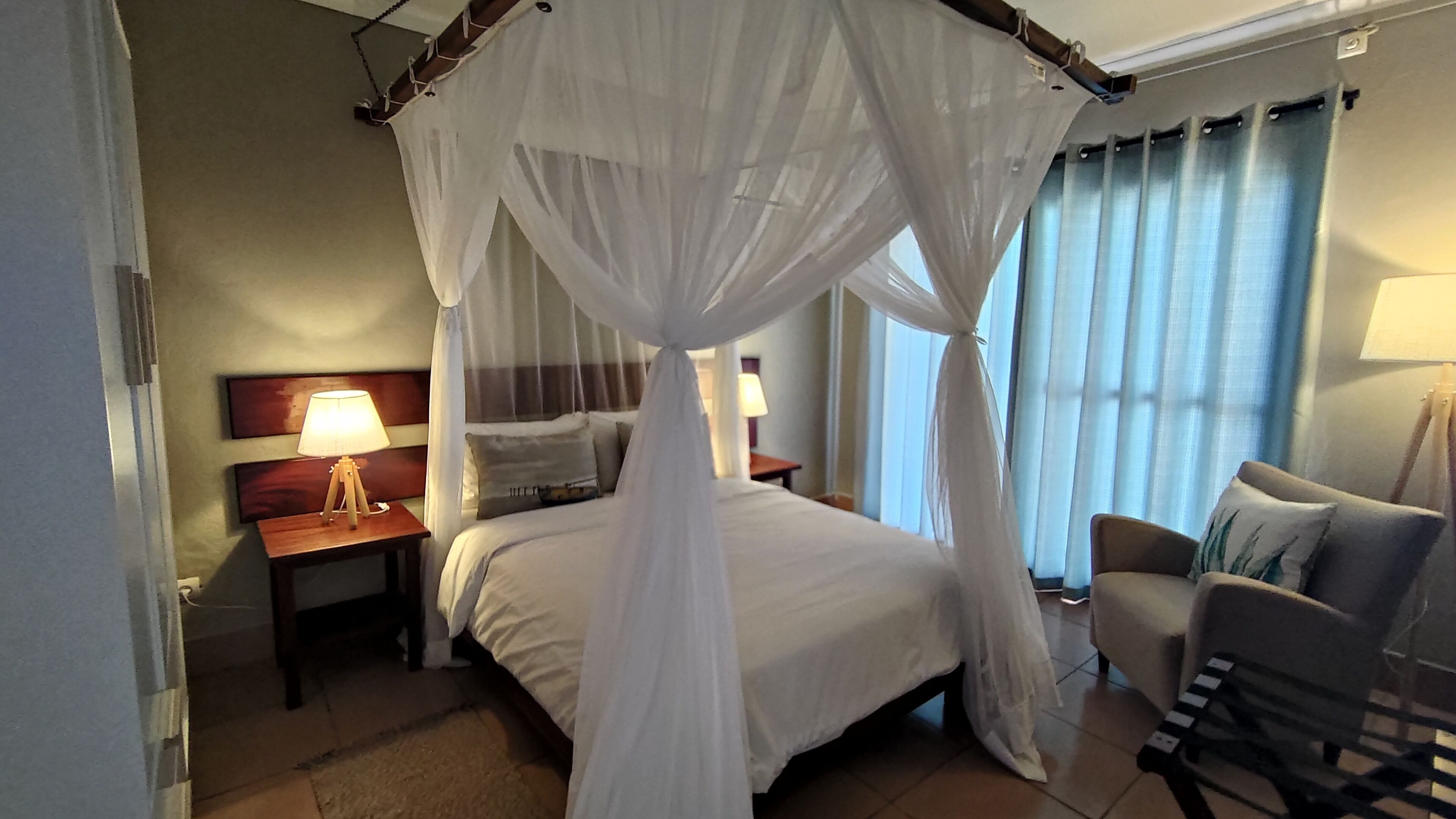 Habitación de hotel con una cama bajo un dosel blanco, una mesita de noche con lámpara, una silla y vistas al exterior a través de cortinas.
