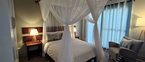 Habitación de hotel con una cama bajo un dosel blanco, una mesita de noche con lámpara, una silla y vistas al exterior a través de cortinas.