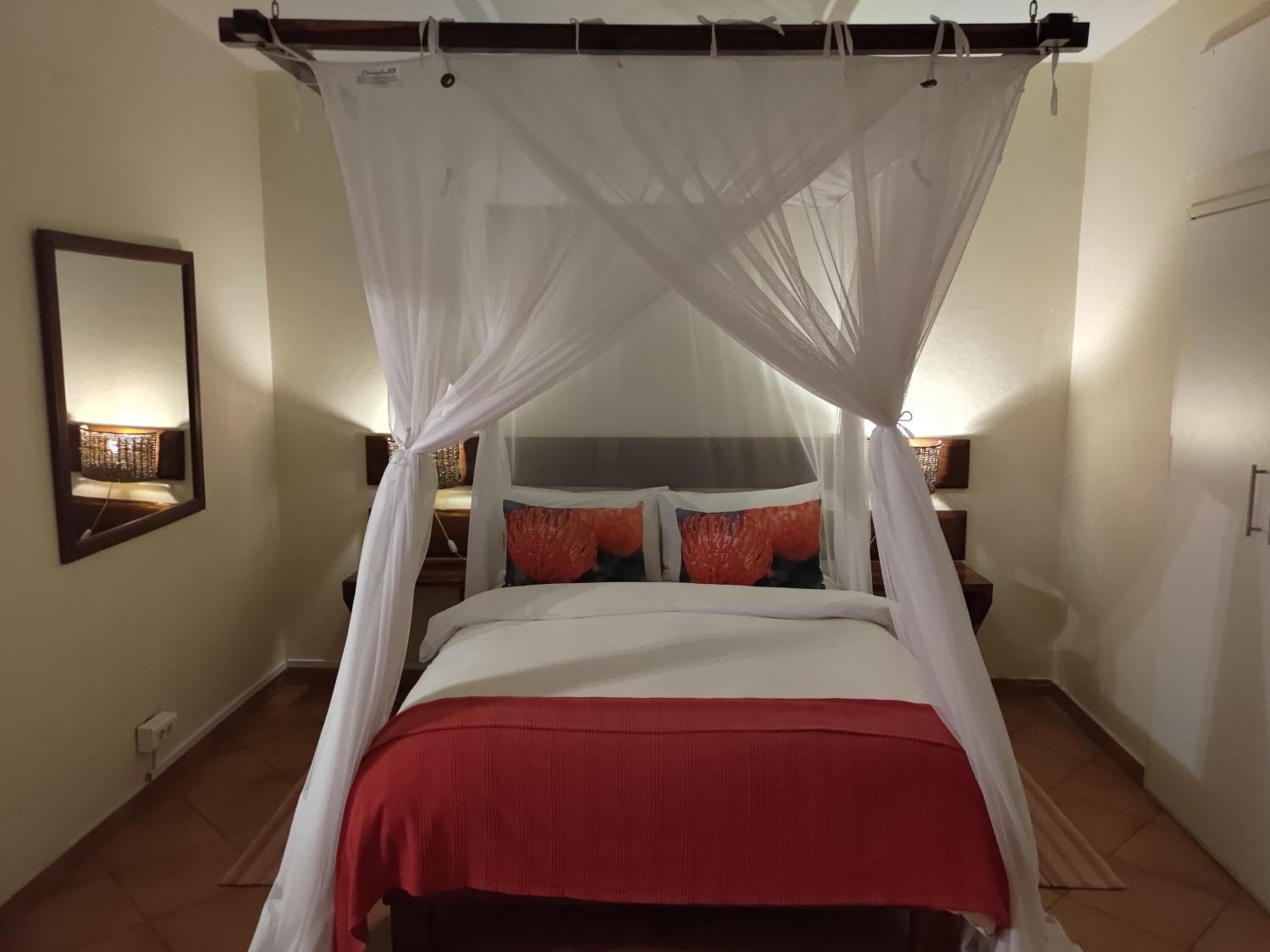 A bedroom with a four-poster canopy bed, a bedside table with a lamp, and a wall-mounted mirror.