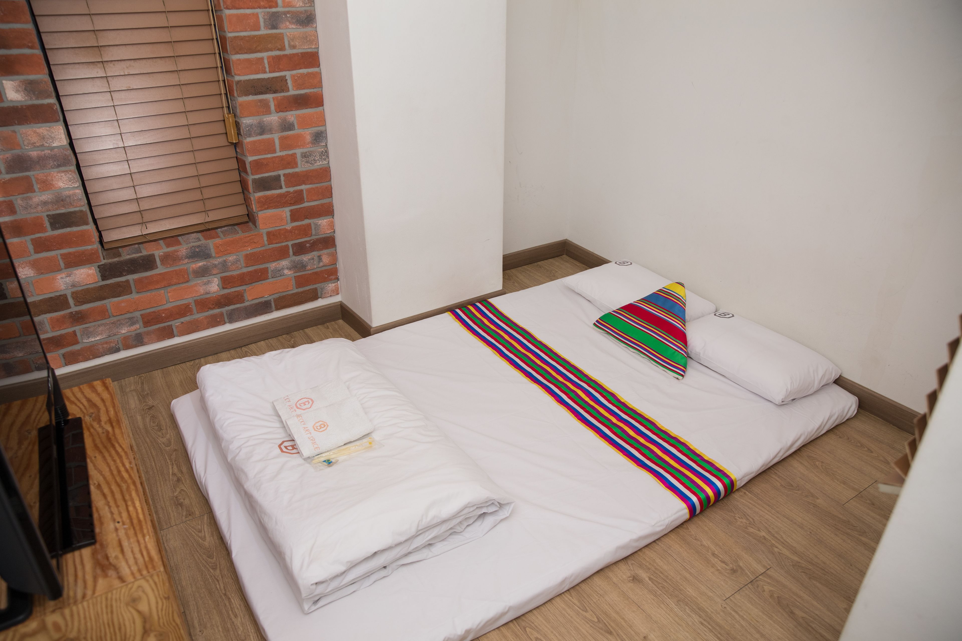 Una cama pequeña y bien hecha con ropa blanca y una manta a rayas coloridas, una almohada blanca y una toalla blanca doblada en el suelo.