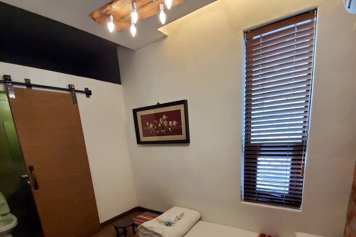 A small, neatly arranged room with a bed, a window with blinds, a framed picture on the wall, and a small table with a colorful cushion.