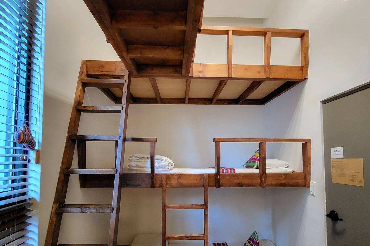 A bunk bed room with a wooden ladder, a small table, and a window with blinds.