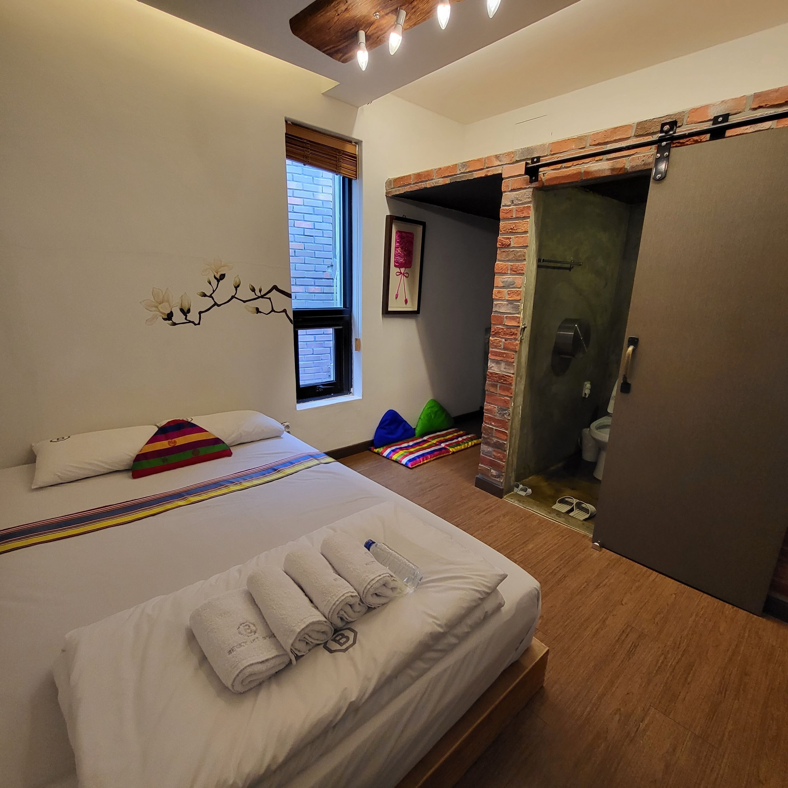 A modern hotel room with a large bed, a window with blinds, a brick fireplace, and a door leading to a bathroom.