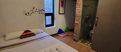 A modern hotel room with a large bed, a window with blinds, a brick fireplace, and a door leading to a bathroom.