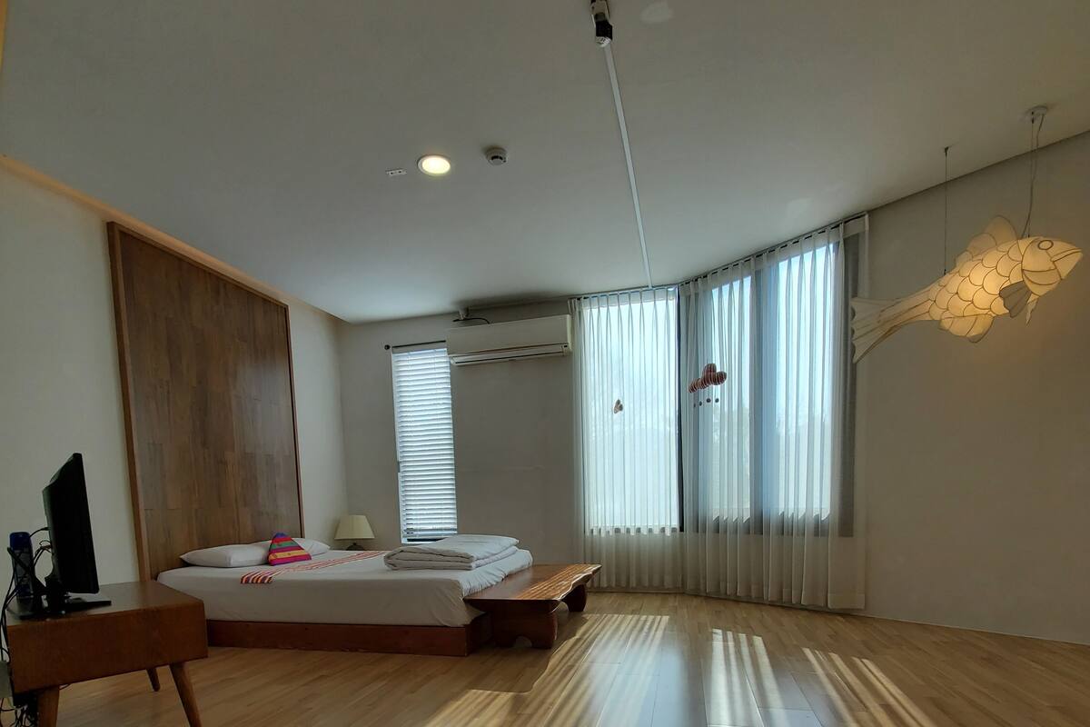 A modern bedroom with a wooden floor, a bed with white bedding, a desk with a computer, and a colorful striped pillow.
