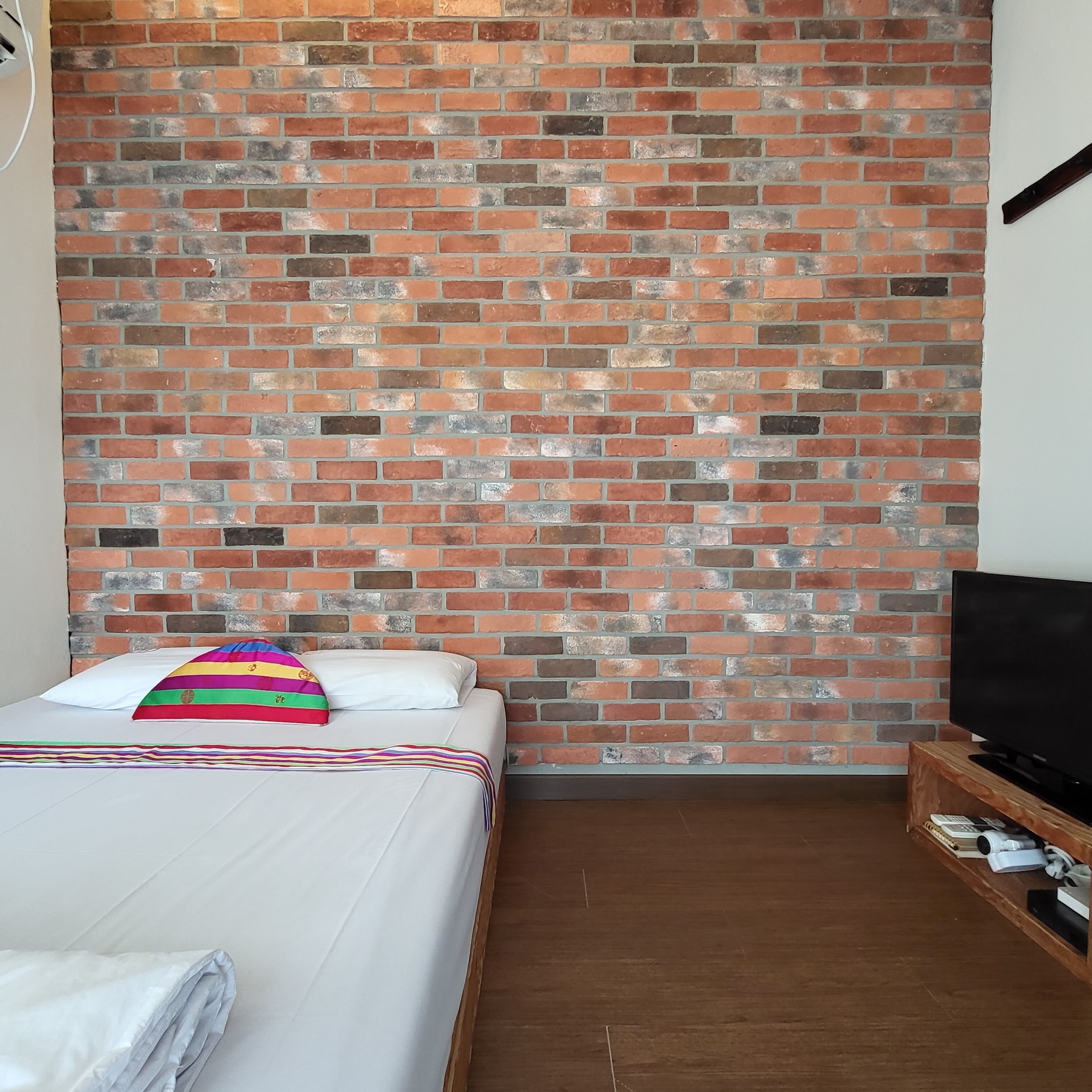 Una habitación de hotel con pared de ladrillo, una cama con ropa blanca y un cojín de colores, y un televisor sobre un mueble de madera.