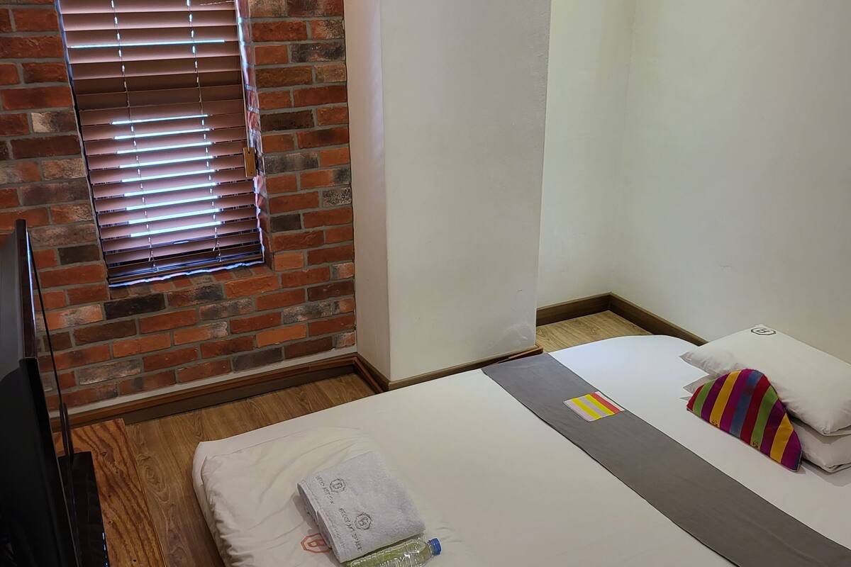 A hotel room with a brick wall, a bed with a white mattress and gray blanket, a colorful striped pillow, and a window with blinds.