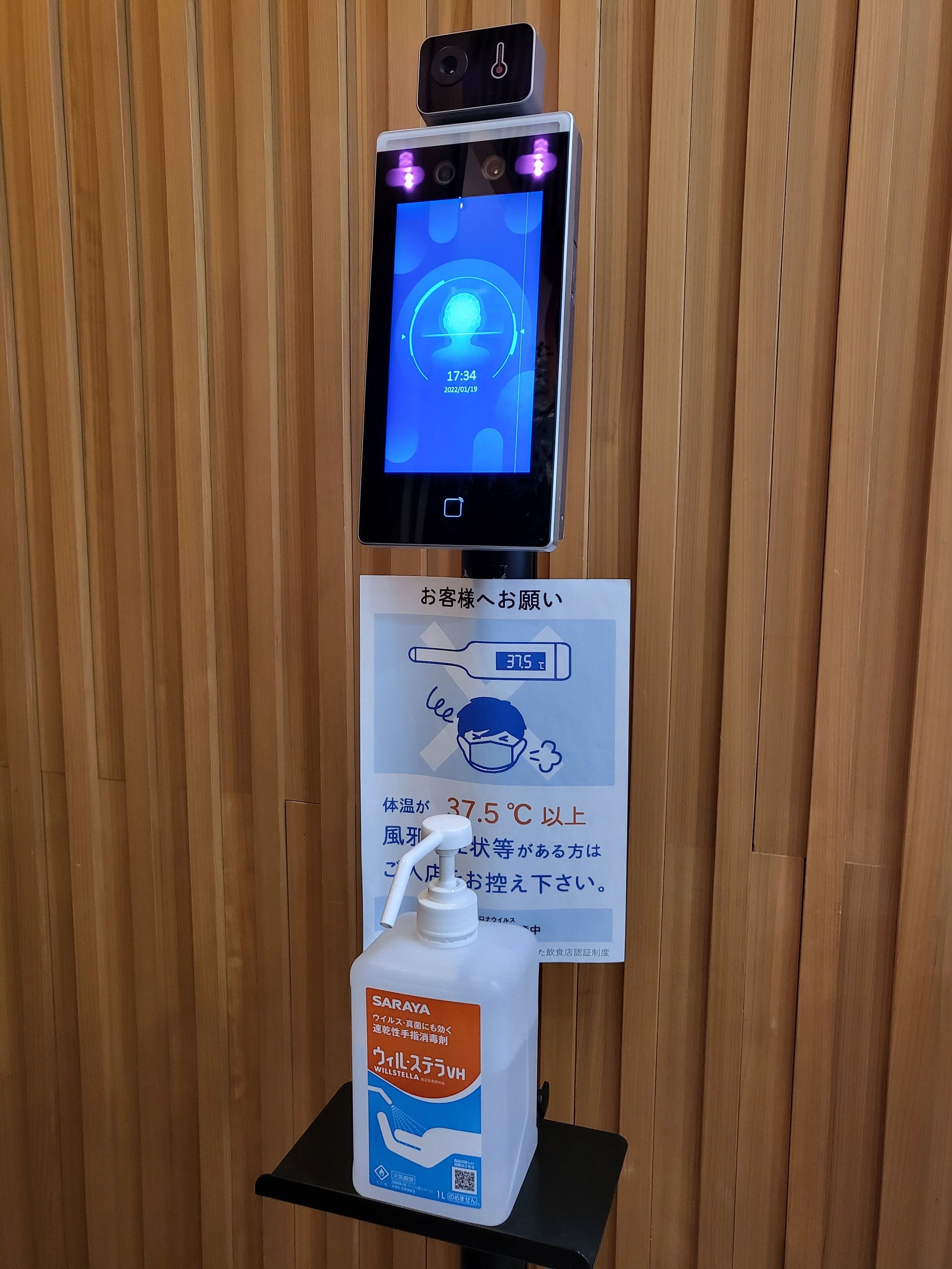 A digital thermometer displaying 37.5°C, a sign instructing to dispose of masks if the temperature is above this, and a sanitizer dispenser.