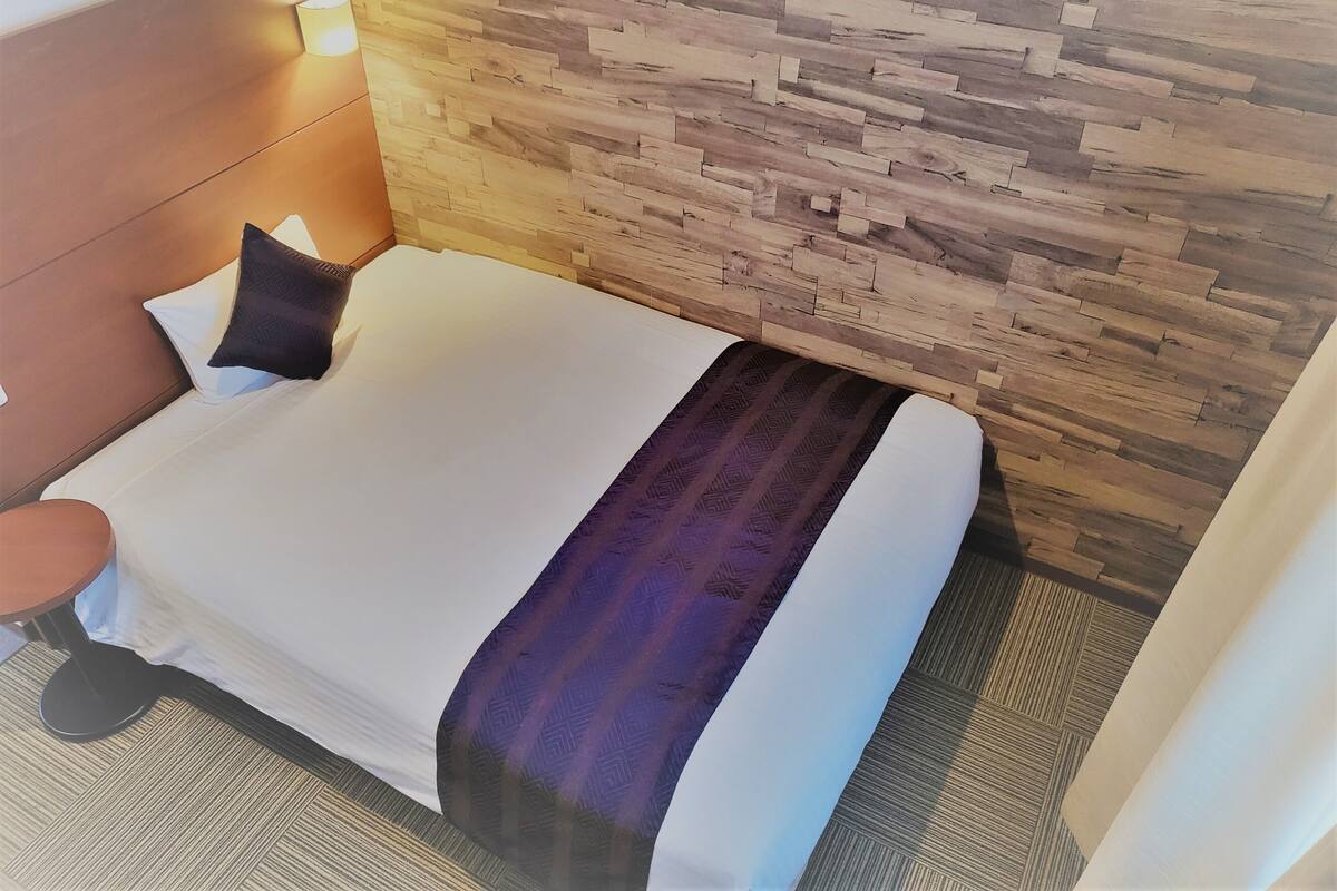 Ein Hotelzimmer mit Holz-Kopfteil, einem Bett mit weißer und violetter Bettwäsche und einem runden Tisch mit einem Stuhl.