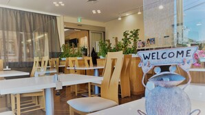 Een restaurant interieur met houten stoelen en tafels, een welkomstbord en potplanten.