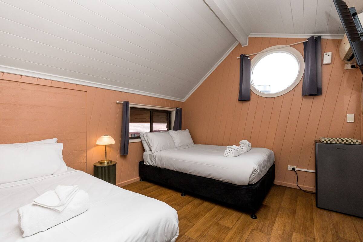 A bedroom with two beds, a small table with a lamp, a round window, and a built-in heater.