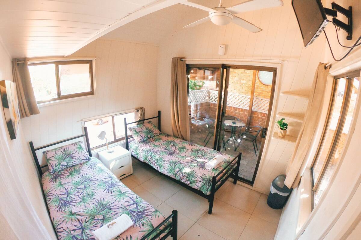 A compact room with two beds, a ceiling fan, a small table, and a sliding glass door leading to an outdoor area.