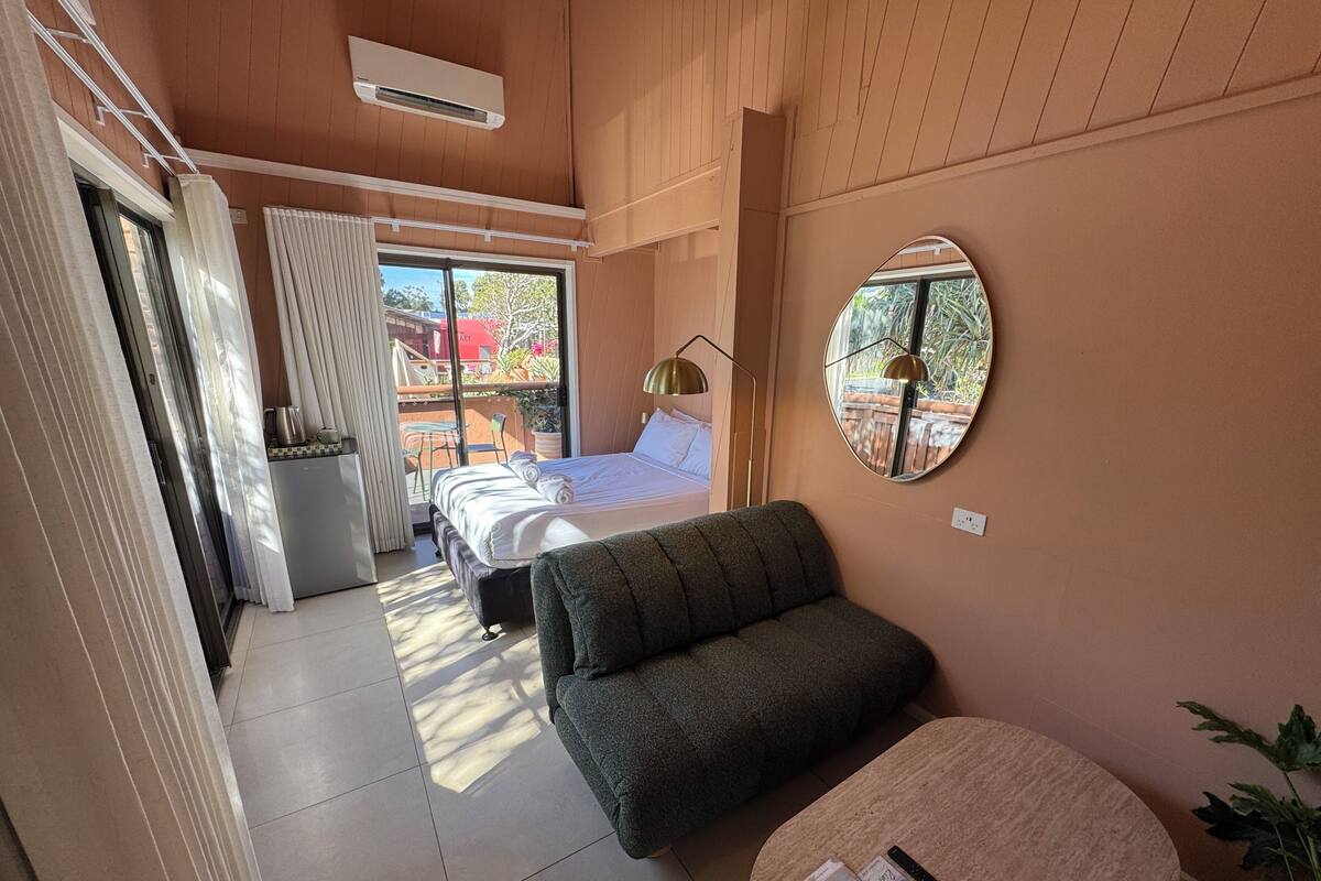 A room with a bed, a sofa, a small table with magazines, a mirror, and a balcony with a view of the outside.