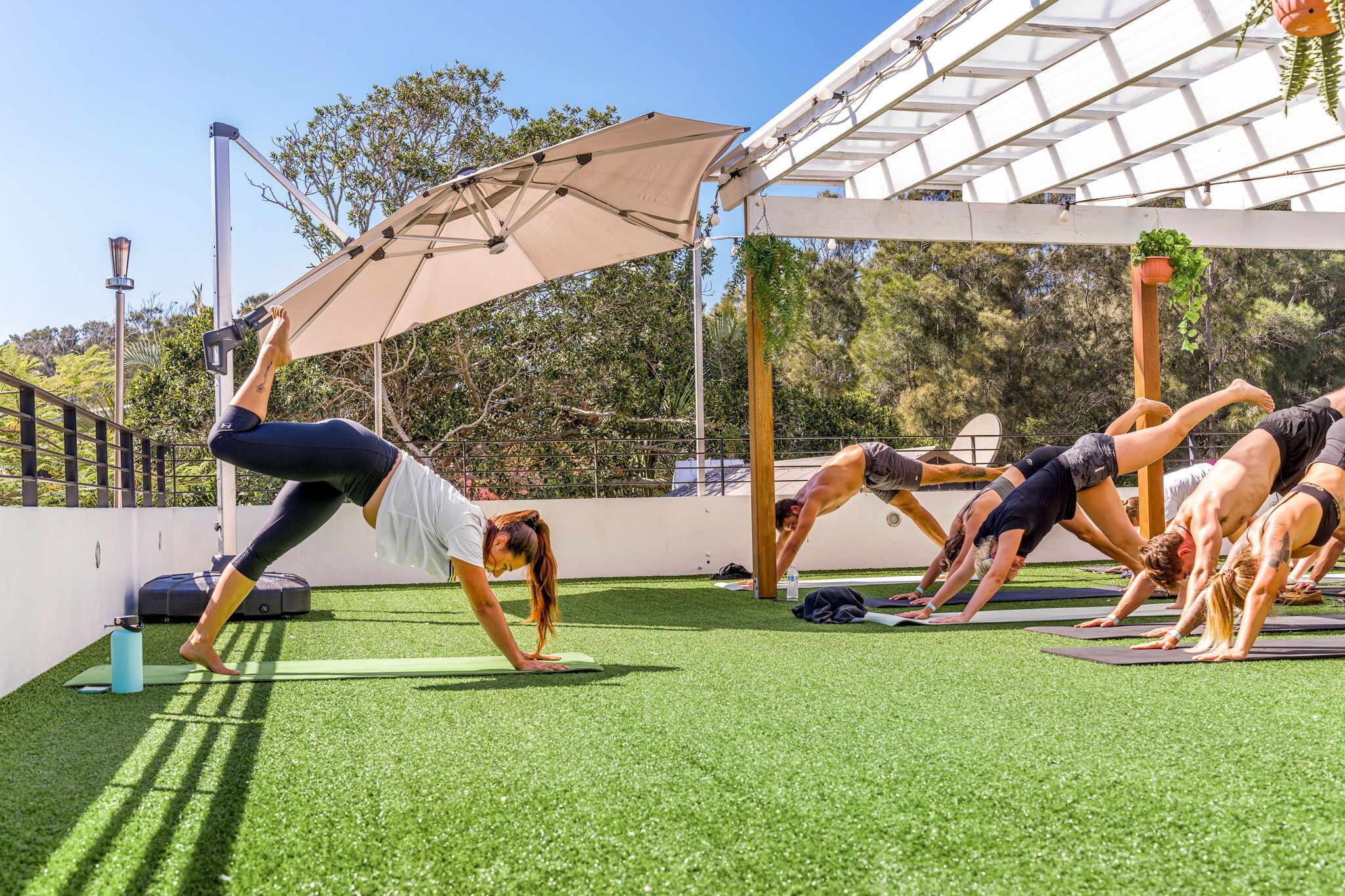 Un grupo de personas practicando yoga en un área al aire libre con césped artificial y una pérgola encima.