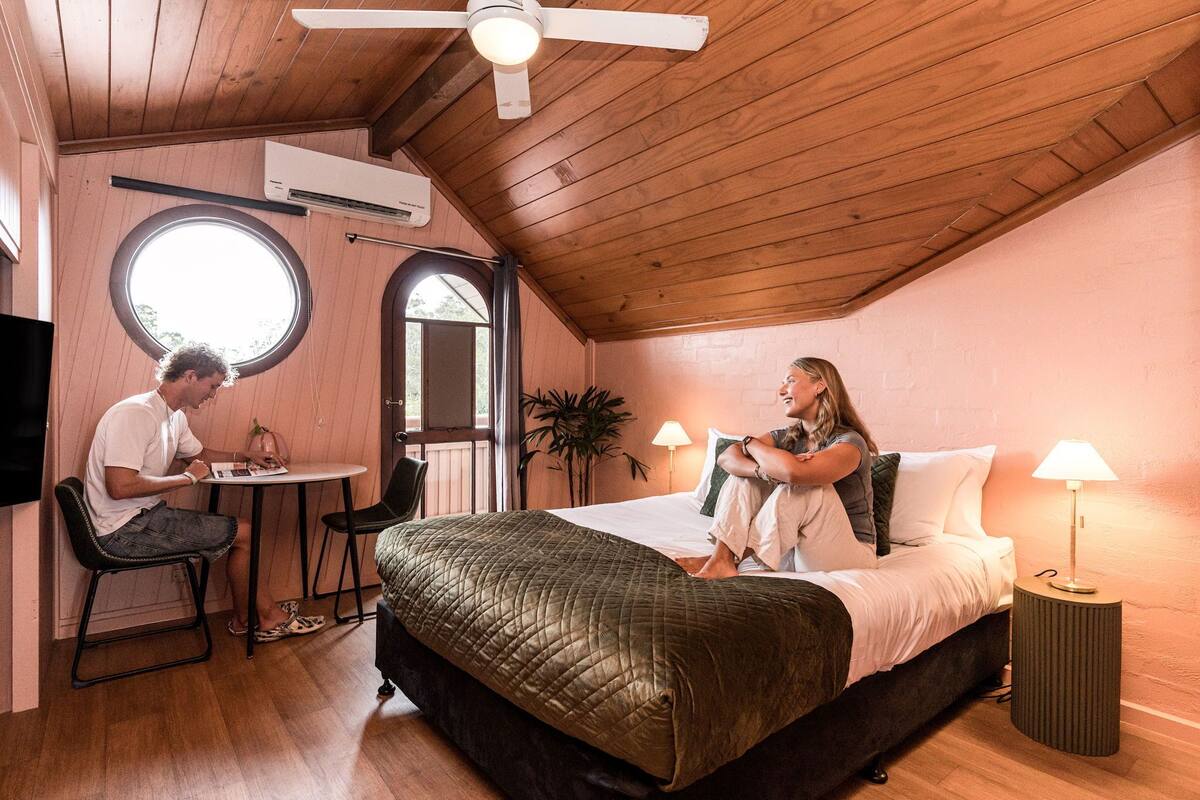 A cozy room with a bed, a round window, a ceiling fan, and a small table with chairs.