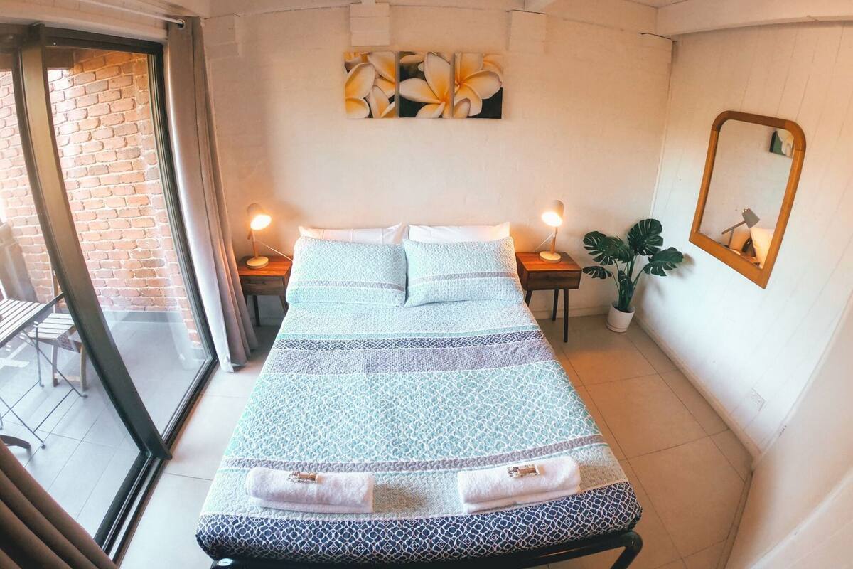 A bedroom with a bed, two bedside tables with lamps, a potted plant, a mirror, and a window with a view of a brick wall.