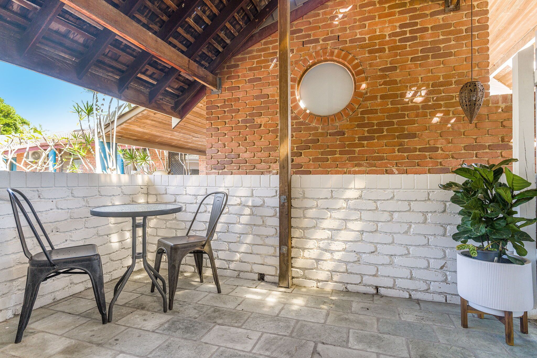 A patio area with two metal chairs and a small round table, a potted plant, and a brick wall with a circular mirror.