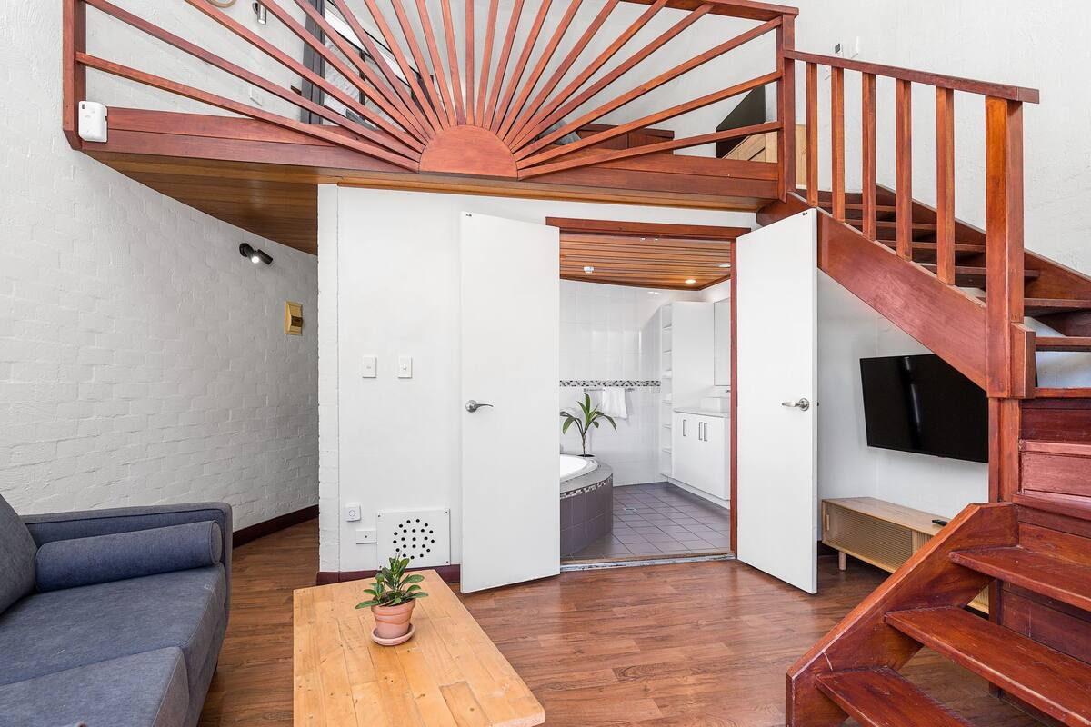 A modern living space with a wooden staircase, a grey sofa, a wooden coffee table with a plant, and an open door leading to a bathroom.