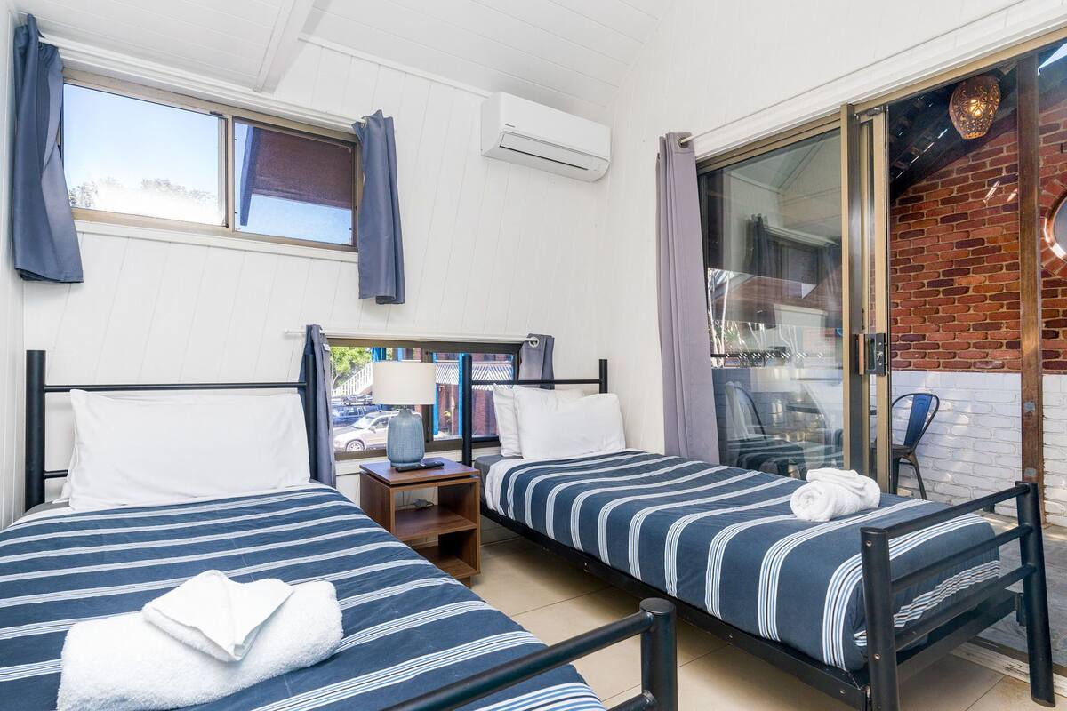 A room with two beds, a window, an air conditioner, and a sliding glass door leading to an outdoor area.