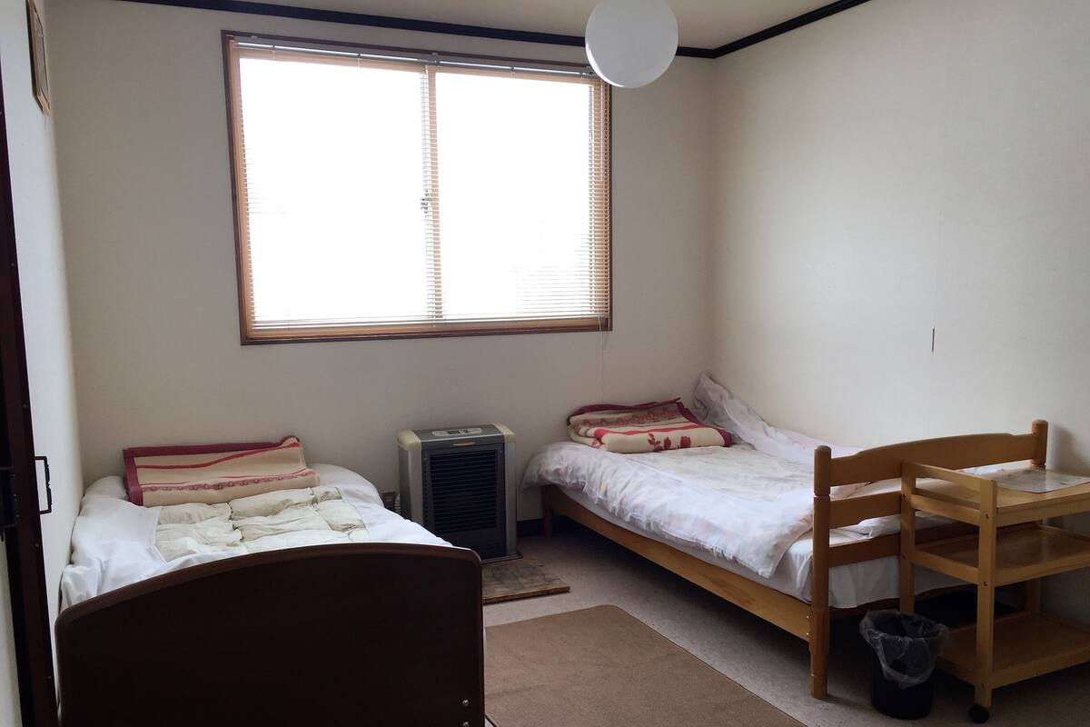 Un dormitorio con dos camas, un calentador, una silla de madera y una ventana con persianas.