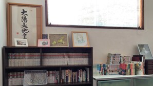 Una biblioteca con estanterías llenas de libros, obras de arte enmarcadas en la pared y una ventana con persianas.