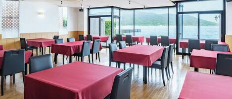 A restaurant with tables covered in red tablecloths, black chairs, and large windows offering a view of a lake and mountains.
