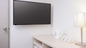 Moderni hotellihuone, jossa on seinälle asennettu taulutelevisio, yöpöytä lampun kanssa, vedenkeitin sekä tarjotinalusta kuppeineen ja ruokalistoineen.